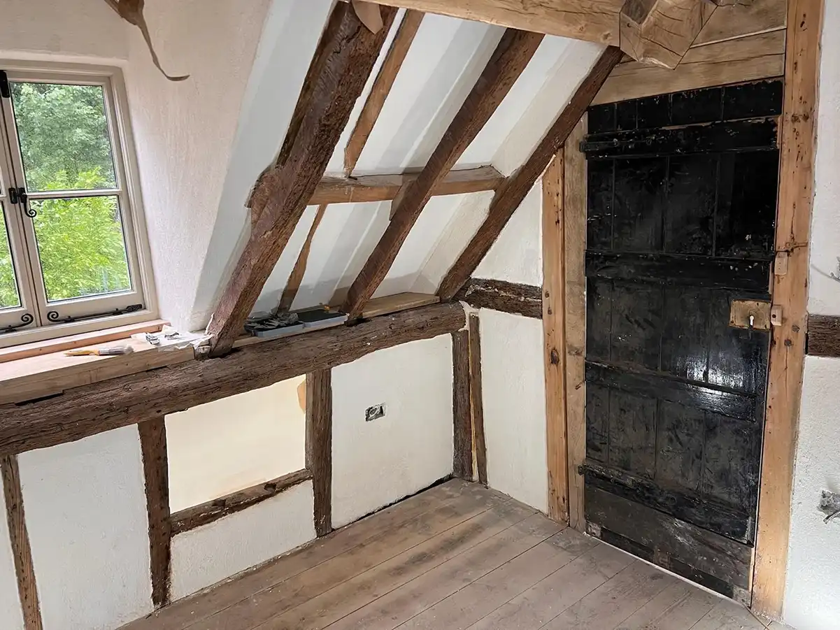Interior of an attic room with wooden beams, a black wooden door, and a window showing green trees outside.
