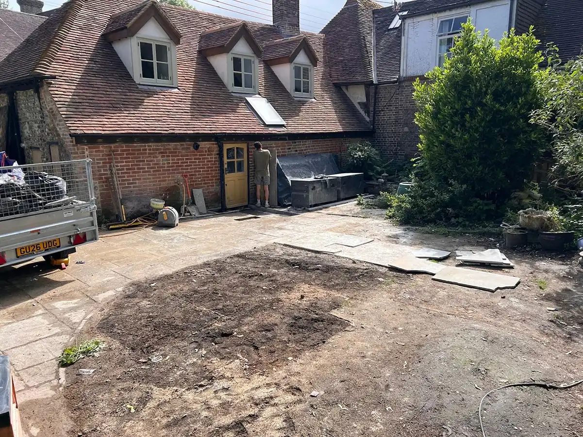 Person working near yellow door of brick house with tiled roof in yard with dirt and stone slabs.